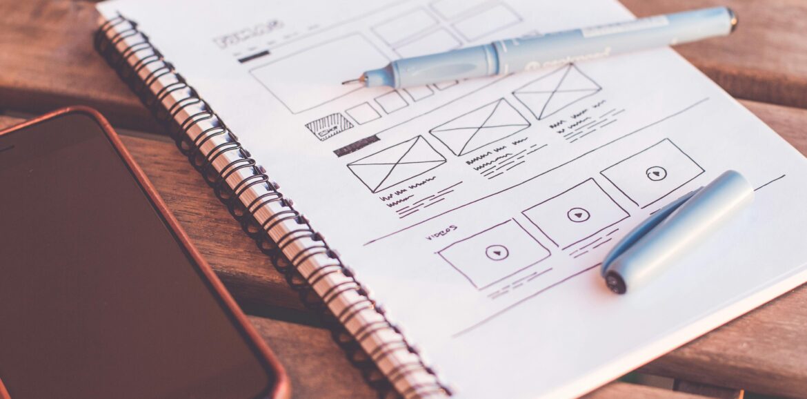 Close-up of a notebook with wireframe sketches and a smartphone on a wooden desk.