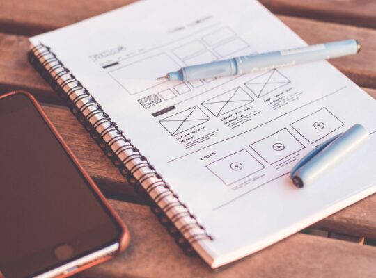 Close-up of a notebook with wireframe sketches and a smartphone on a wooden desk.